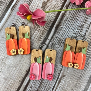 Puede incluir: Tres pares de pendientes de calabaza de madera. Los pendientes est&aacute;n pintados en naranja, rosa y marr&oacute;n. Cada par presenta dos calabazas con una peque&ntilde;a flor en la parte inferior.