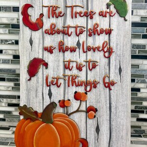 K&ouml;nnte beinhalten: Ein Holzschild mit wei&szlig; get&uuml;nchtem Hintergrund und einer herbstlichen Botschaft. Das Schild lautet "The trees are about to show us how lovely it is to let things go". Das Schild ist mit einem K&uuml;rbis, Bl&auml;ttern und Beeren verziert.
