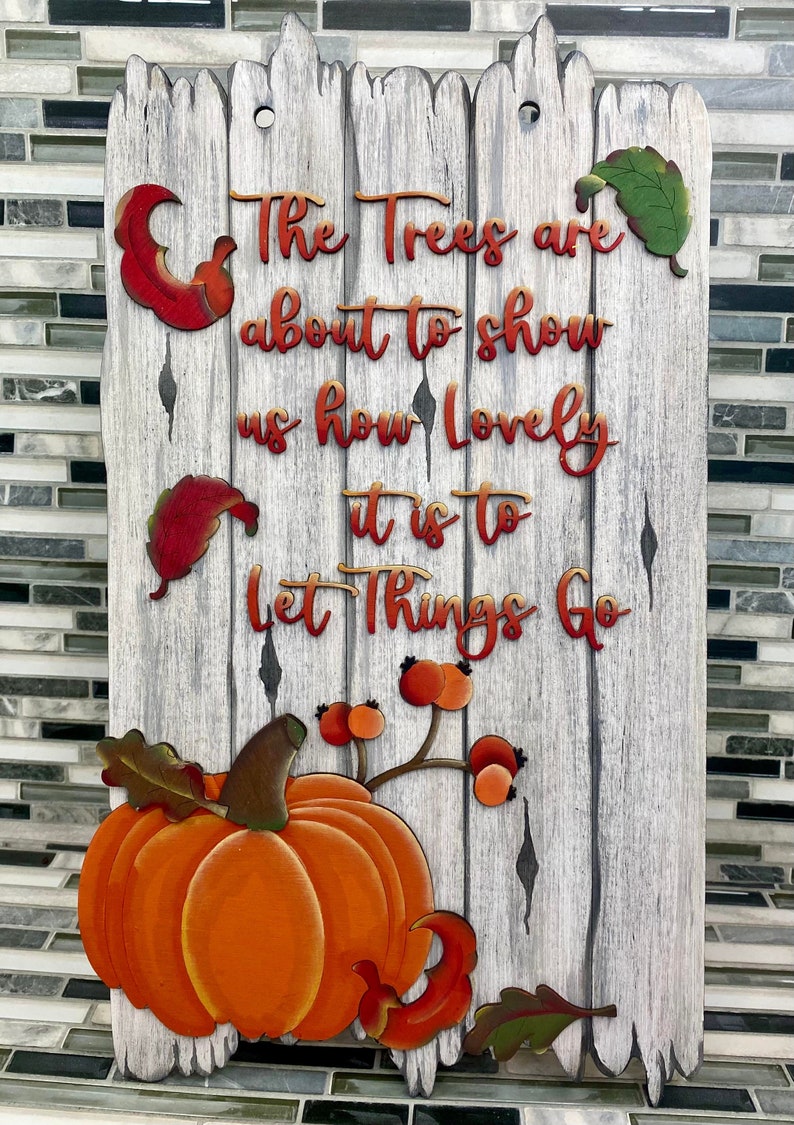 K&ouml;nnte beinhalten: Ein Holzschild mit wei&szlig; get&uuml;nchtem Hintergrund und einer herbstlichen Botschaft. Das Schild lautet "The trees are about to show us how lovely it is to let things go". Das Schild ist mit einem K&uuml;rbis, Bl&auml;ttern und Beeren verziert.