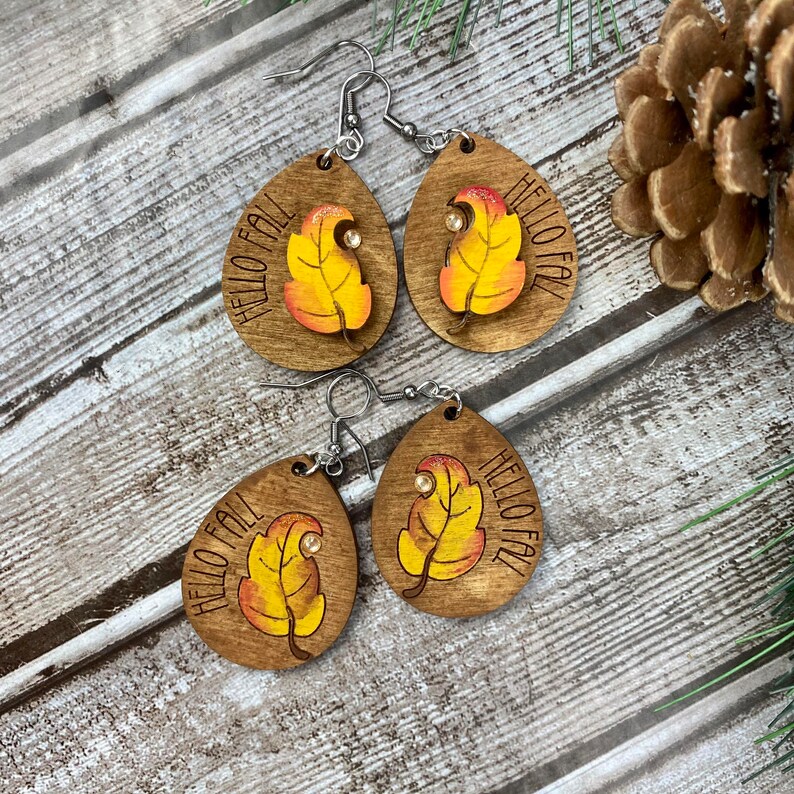 May include: A pair of wooden earrings with a teardrop shape. Each earring features a painted leaf with orange, yellow, and red colors and the text "Hello Fall".