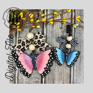 Pode incluir: Dois enfeites de borboleta com bolinhas pretas e brancas. Uma borboleta é rosa e a outra é azul. Cada borboleta tem uma conta de madeira e um laço. O laço da borboleta rosa é de estampa de leopardo e o laço da borboleta azul é xadrez preto e branco.