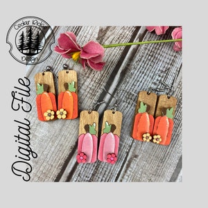 Puede incluir: Un par de pendientes de madera con un dise&ntilde;o de calabaza. Las calabazas est&aacute;n pintadas de naranja y rosa con tallos verdes y flores amarillas. Los pendientes est&aacute;n unidos a ganchos plateados.