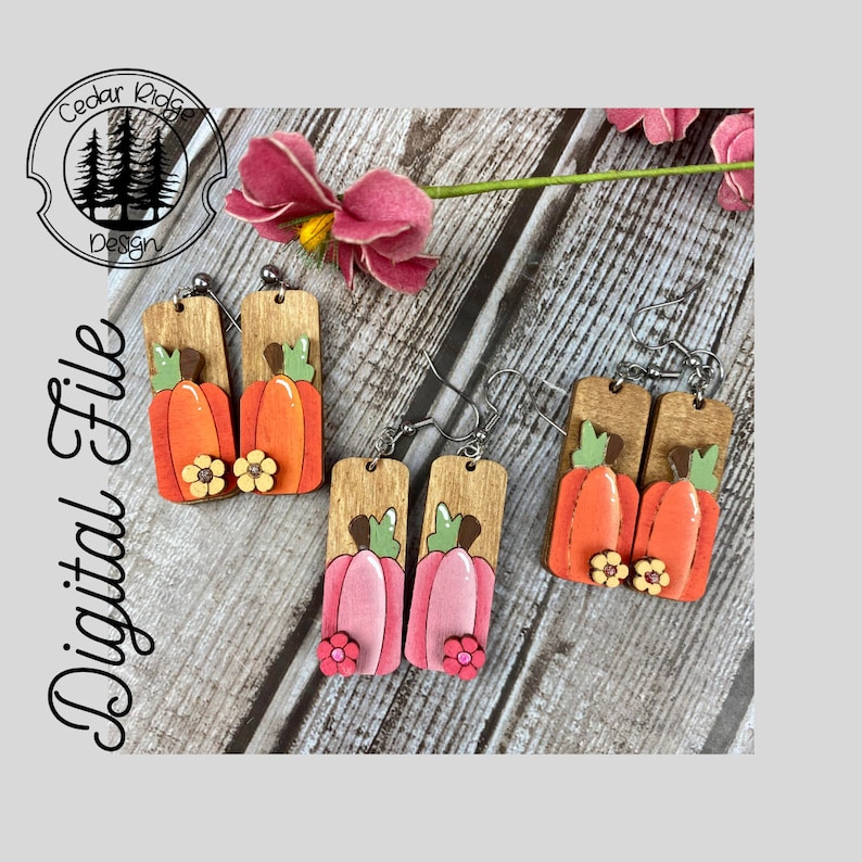 Puede incluir: Un par de pendientes de madera con un dise&ntilde;o de calabaza. Las calabazas est&aacute;n pintadas de naranja y rosa con tallos verdes y flores amarillas. Los pendientes est&aacute;n unidos a ganchos plateados.