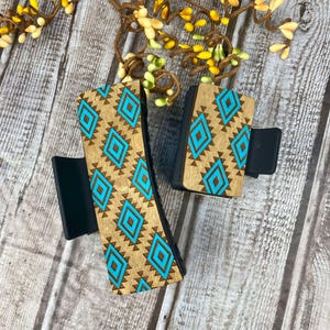 May include: Two wooden hair clips with a turquoise and brown geometric pattern. The clips are made of wood and have a black plastic base.