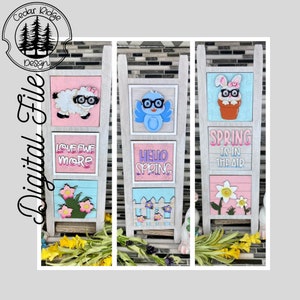 May include: Three wooden spring-themed tiered trays with different designs. The top tier of the first tray features a white lamb with glasses, the second tier says "Love ewe more", and the third tier has pink flowers. The top tier of the second tray features a blue bird with glasses, the second tier says "Hello Spring", and the third tier has a white picket fence. The top tier of the third tray features a white bunny with glasses in a pot, the second tier says "Spring is in the air", and the third tier has a white flower.