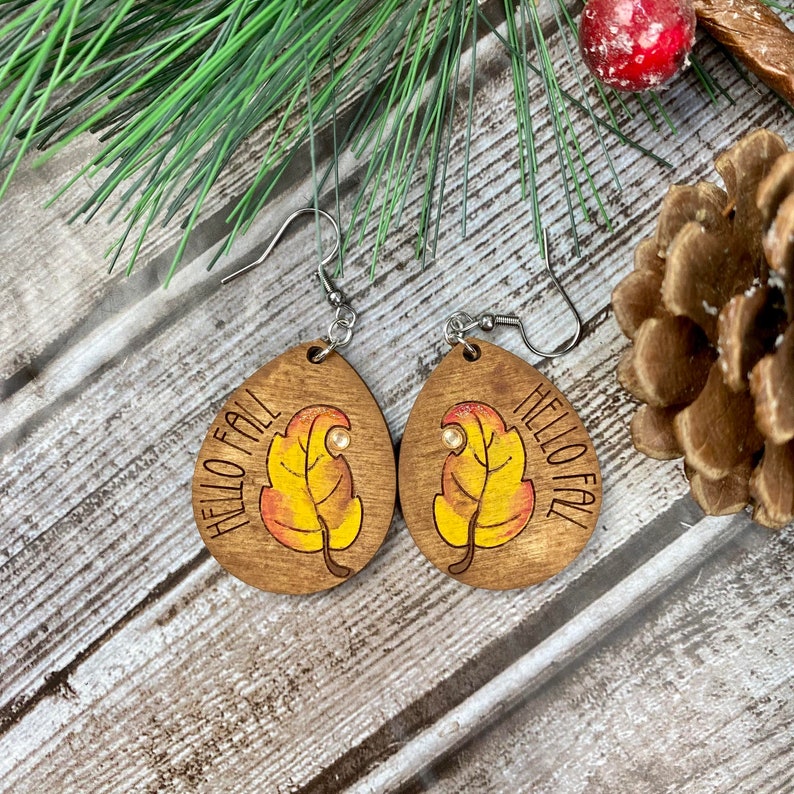 May include: A pair of wooden earrings with a fall leaf design and the text "Hello Fall". The earrings are brown with yellow and orange leaves and a silver hook.
