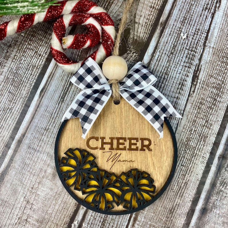 Op de afbeelding: Houten kerstversiering met een zwart-wit geblokte strik, een zwart-geel juichontwerp en de tekst "Cheer Mama".