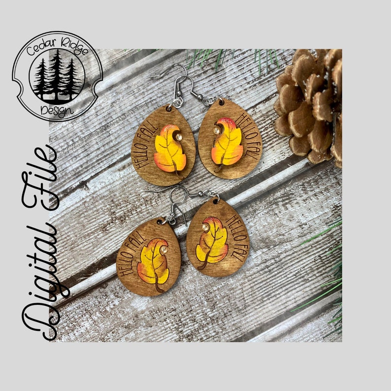 May include: A pair of wooden earrings with a teardrop shape. The earrings are painted with a yellow and orange leaf design and the words "Hello Fall".