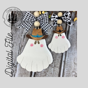 Puede incluir: Dos adornos de fantasmas blancos con sombreros de vaquero marrones y mejillas rosadas. El fantasma más grande tiene una joya azul en su sombrero. Los fantasmas están colgados de una superficie de madera con una cinta a cuadros en blanco y negro.