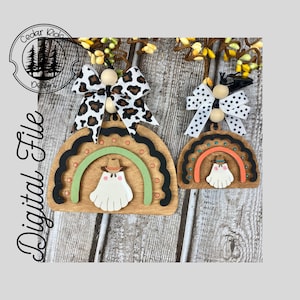 Può includere: Due ornamenti in legno a forma di arcobaleno con un fantasma che indossa un cappello da cowboy al centro. L'ornamento più grande ha un fiocco con stampa leopardata bianco e nero e l'ornamento più piccolo ha un fiocco con pois bianco e nero.