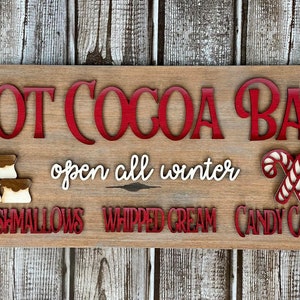 K&ouml;nnte beinhalten: Ein Holzschild mit dem Text "Hot Cocoa Bar" in roten Buchstaben. Das Schild enth&auml;lt auch den Text "open all winter" und listet die Toppings auf: "marshmallows", "whipped cream" und "candy canes". Das Schild ist mit zwei Tassen hei&szlig;er Schokolade mit Marshmallows verziert, die mit einem rot-wei&szlig; gestreiften Strohhalm und einer roten Schleife gekr&ouml;nt sind.