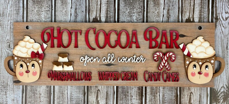 K&ouml;nnte beinhalten: Ein Holzschild mit dem Text "Hot Cocoa Bar" in roten Buchstaben. Das Schild enth&auml;lt auch den Text "open all winter" und listet die Toppings auf: "marshmallows", "whipped cream" und "candy canes". Das Schild ist mit zwei Tassen hei&szlig;er Schokolade mit Marshmallows verziert, die mit einem rot-wei&szlig; gestreiften Strohhalm und einer roten Schleife gekr&ouml;nt sind.