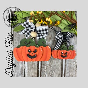 Könnte beinhalten: Zwei hölzerne Kürbisdekorationen mit schwarz-weißen Karobändern. Die Kürbisse sind orange lackiert mit schwarzen Kürbisgesichtern. Der größere Kürbis hat drei übereinander gestapelte Kürbisse, während der kleinere Kürbis zwei übereinander gestapelte Kürbisse hat.