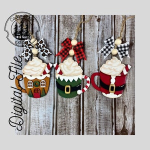 May include: Three wooden Christmas ornaments shaped like mugs of hot chocolate with whipped cream and a candy cane. The mugs are decorated with gingerbread house, elf, and Santa Claus themes. Each mug has a bow and a wooden bead.