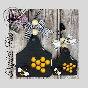 May include: Two black wooden bee-themed ornaments with a honeycomb design and a bee illustration. The ornaments are shaped like a cow tag and have a white ribbon with black polka dots and a white ribbon with black and yellow bees.