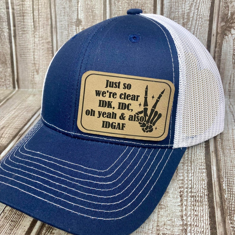 May include: A navy blue and white trucker hat with a brown patch that says "Just so we're clear IDK, IDC, oh yeah & also IDgaf" with a skeleton hand making the peace sign.