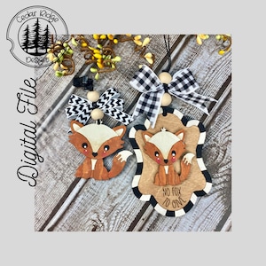 Può includere: Due ornamenti di legno a forma di volpe con nastro a scacchi bianco e nero. L'ornamento più grande ha il testo "No fox to give".