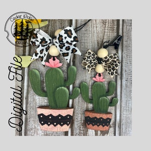 Può includere: Due ornamenti di cactus in legno con fiori rosa, vasi marroni con pois neri e bianchi e fiocchi leopardati con perline di legno. Gli ornamenti sono appesi a un cordoncino nero e una clip.