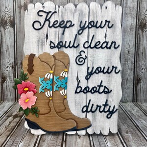 May include: Wooden sign with a whitewashed background and black lettering that reads "Keep your soul clean & your boots dirty". The sign features two brown cowboy boots with turquoise and white floral designs and pink flowers.