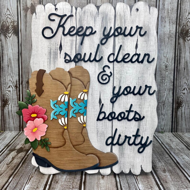 May include: Wooden sign with a whitewashed background and black lettering that reads "Keep your soul clean & your boots dirty". The sign features two brown cowboy boots with turquoise and white floral designs and pink flowers.