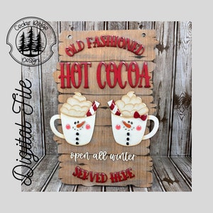 May include: A wooden sign with the text "Old Fashioned Hot Cocoa" in red letters. Two white mugs with snowman faces and red bows are on either side of the text. The sign also says "open all winter" and "Served Here".