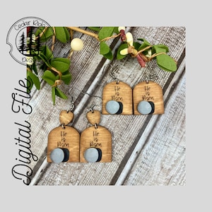 May include: A pair of wooden earrings with a heart-shaped top and a rounded bottom. The bottom of the earrings feature a gray circle with the text "He is Risen".
