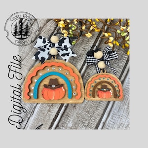 Könnte beinhalten: Zwei hölzerne Kürbis-Regenbogen-Ornamente mit einem Cowboyhut auf dem Kürbis. Das größere Ornament hat einen türkisfarbenen Regenbogen mit Strasssteinen und das kleinere Ornament hat einen braunen Regenbogen mit weißen Punkten. Beide Ornamente haben einen braunen Holzgrund.