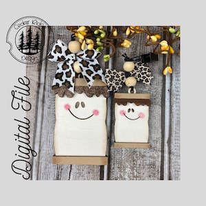 May include: Two wooden cutout ornaments shaped like marshmallows with smiling faces. The ornaments are decorated with brown and white faux fur trim and have a brown wooden base. Each ornament has a brown and white leopard print bow with a wooden bead accent.
