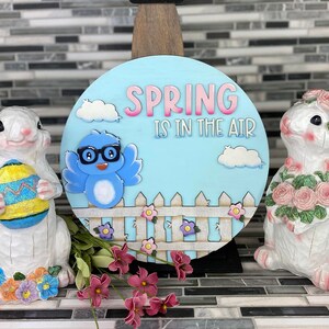 May include: A blue and white wooden sign with the text "Spring is in the air" and a cartoon blue bird wearing glasses. The sign is surrounded by white picket fence and pink flowers. Two white ceramic bunny figurines are on either side of the sign, one holding a pink flower bouquet and the other holding a yellow and blue Easter egg.