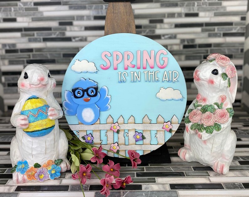 May include: A blue and white wooden sign with the text "Spring is in the air" and a cartoon blue bird wearing glasses. The sign is surrounded by white picket fence and pink flowers. Two white ceramic bunny figurines are on either side of the sign, one holding a pink flower bouquet and the other holding a yellow and blue Easter egg.