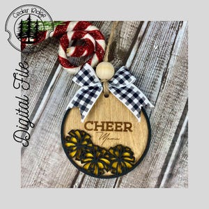 Op de afbeelding: Een ronde houten ornament met een zwart-wit geblokte strik. De ornament heeft het woord "CHEER" en het woord "Mama" erop geschreven. De ornament is versierd met gele en zwarte pompoenen.