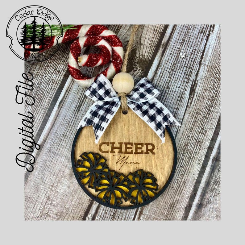 Op de afbeelding: Een ronde houten ornament met een zwart-wit geblokte strik. De ornament heeft het woord "CHEER" en het woord "Mama" erop geschreven. De ornament is versierd met gele en zwarte pompoenen.