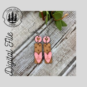 Puede incluir: Un par de pendientes de madera con corazones rosas y el texto "Be Mine" en cada pendiente.