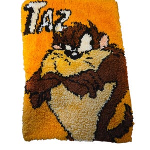 Puede incluir: Alfombra rectangular hecha a mano con el personaje de dibujos animados Taz. La alfombra tiene un fondo amarillo con la palabra "TAZ" en blanco. El Demonio de Tasmania está representado en tonos marrones, beige y blancos.