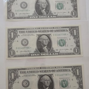 May include: Three consecutive United States one-dollar Federal Reserve notes, each featuring a portrait of George Washington, the text Federal Reserve Note, The United States of America, and One Dollar, with serial numbers D01487785, D01487786, and D01487787.