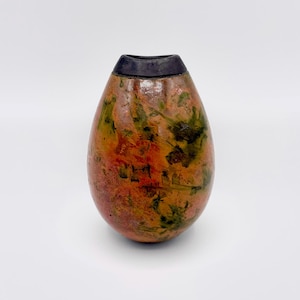 Könnte beinhalten: Eine kleine, eiförmige Vase mit einem glatten, abgerundeten Körper, der ein gesprenkeltes Muster in Orange-, Gelb- und Dunkelgrüntönen aufweist. Die Vase hat eine schmale, kreisförmige Öffnung an der Oberseite, die mit einem soliden, mattschwarzen Rand versehen ist, der einen Kontrast zur lebendigen, mehrfarbigen Oberfläche des Hauptkörpers bildet.