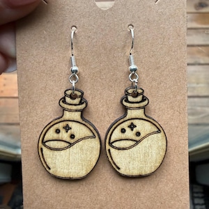 May include: A pair of wooden earrings featuring a potion bottle design. The earrings are laser-cut from light-colored wood and have a silver-colored hook. Each bottle has a curved liquid line and star details.