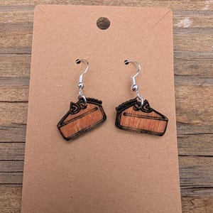 May include: A pair of wooden earrings shaped like slices of pie. The earrings have a silver hook and are made of light brown wood with a darker brown outline. The pie slice has a bow on top.