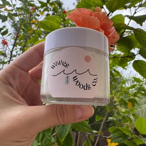 May include: A clear glass jar with a white lid is held against a backdrop of green foliage and a peach-coloured flower. The jar features a circular label with the text "waves woods co." and a minimalist design of waves and a tree.