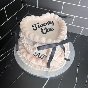 Könnte beinhalten: Zweistöckige herzförmige Torte mit hellrosa Zuckerguss und dekorativen Verzierungen. Die obere Schicht trägt die Aufschrift "Twenty One" in schwarzer Schrift, die untere Schicht die Initialen "AJ". Schwarze Bänder schmücken die Torte.