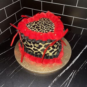 Può includere: Una torta a forma di cuore con una parte superiore con stampa leopardata e bordi di glassa rossa. La torta ha un bordo di glassa nero ed è decorata con nastri rossi e paillettes dorate. Si trova su un vassoio dorato.