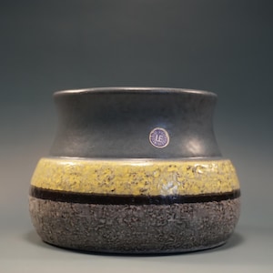 May include: A ceramic vase with a textured, grey base, a yellow band, and a dark grey top. A thin black stripe separates the yellow and grey sections. The vase has a small, circular sticker on the front.