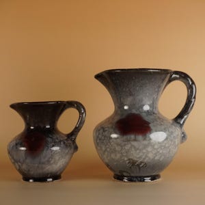 Puede incluir: Dos jarras de cerámica con un esmalte moteado gris y negro y un acento rojo oscuro. Las jarras tienen un asa y un pico. La jarra más grande mide aproximadamente 20 cm de altura.