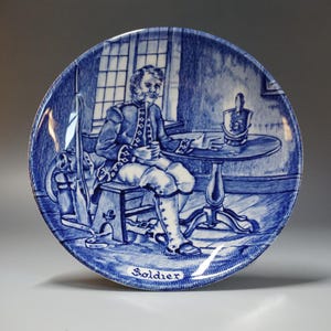 May include: A blue and white decorative plate featuring a detailed illustration of a soldier seated at a table. The plate has a classic design with the word "Soldier" at the bottom. The scene includes a rifle and a window.
