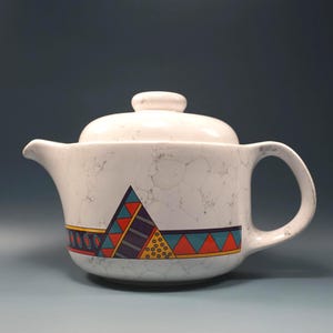 May include: White ceramic teapot with lid. The teapot features a geometric design in red, orange, teal, and purple. The teapot has a handle and a spout. The surface has a crackle pattern.