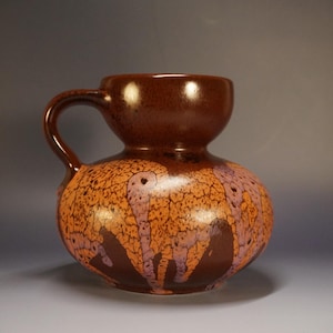 May include: A brown ceramic jug with a handle and a rounded body. The jug features a textured orange and purple pattern over a dark brown base. The top is a darker shade of brown.