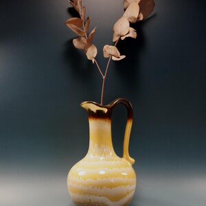 May include: A ceramic pitcher with a yellow and brown glaze, featuring horizontal stripes. The pitcher has a handle and a spout, and is filled with dried eucalyptus branches. The background is a dark teal color.