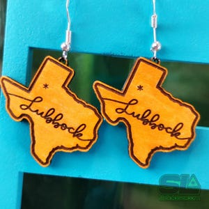 May include: A pair of wooden earrings in the shape of Texas, with the word "Lubbock" in cursive. The earrings are light orange with a dark brown outline and silver ear wires. They are displayed against a blue background.