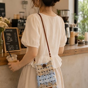 May include: A rectangular crochet crossbody bag featuring a shell stitch pattern in alternating horizontal stripes of light blue, beige, and cream. The bag has a single dark brown wooden button closure and a thin, adjustable brown leather strap.