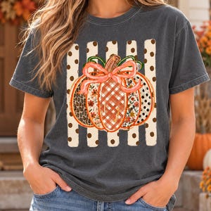 May include: A charcoal gray short-sleeve t-shirt featuring a central graphic of a pumpkin. The pumpkin design incorporates plaid, leopard print, and floral patterns, topped with a coral-colored bow, set against a background of vertical stripes.